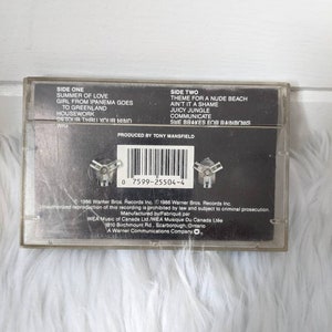 May include: Black cassette tape with white text listing the songs on each side. The text reads "Side One: Summer of Love, Girl from Ipanema Goes to Greenland, Housework, Detour Thru Your Mind, Wig" and "Side Two: Theme for a Nude Beach, Ain't It a Shame, Juicy Jungle, Communicate, She Brakes for Rainbows". The text "Produced by Tony Mansfield" is also included.
