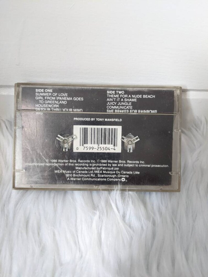 May include: Black cassette tape with white text listing the songs on each side. The text reads "Side One: Summer of Love, Girl from Ipanema Goes to Greenland, Housework, Detour Thru Your Mind, Wig" and "Side Two: Theme for a Nude Beach, Ain't It a Shame, Juicy Jungle, Communicate, She Brakes for Rainbows". The text "Produced by Tony Mansfield" is also included.