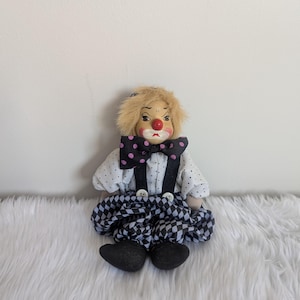Può includere: Un piccolo clown di porcellana con una gonna a scacchi bianchi e neri, una camicia bianca con pois neri, un papillon nero con pois rosa e un cappello bianco e nero. Il clown ha un viso dipinto con labbra rosse, un naso rosso e sopracciglia nere.