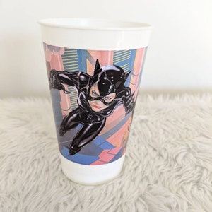 May include: White plastic cup with a colorful image of Catwoman in a black catsuit, leaping through a city scene. The cup is on a white fluffy surface.