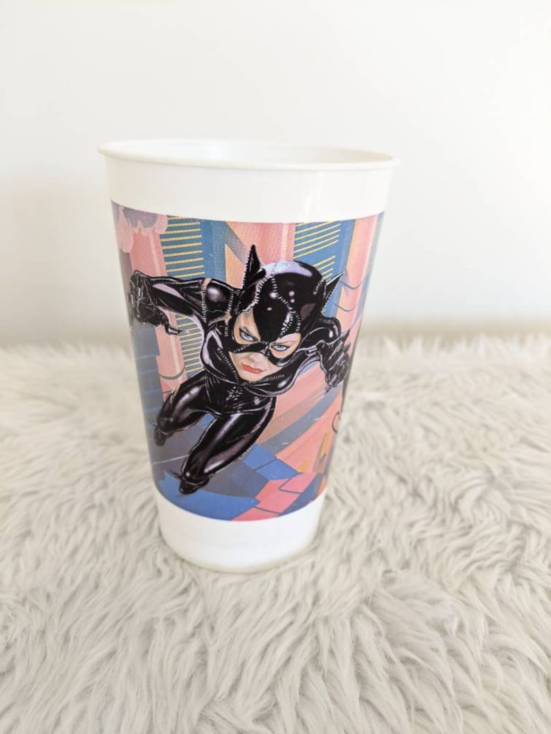 May include: White plastic cup with a colorful image of Catwoman in a black catsuit, leaping through a city scene. The cup is on a white fluffy surface.