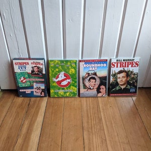 May include: Four DVD cases featuring Bill Murray movies. The cases are arranged from left to right: Stripes Extended Cut, Ghostbusters, Groundhog Day, and Stripes. The cases are all in good condition and have a similar design.