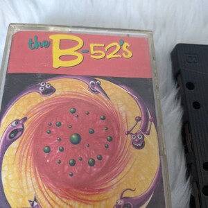May include: A cassette tape featuring a red, yellow and purple illustration of a swirling galaxy with cartoon worm-like creatures. The text "the B-52's" is printed in yellow on a red background.
