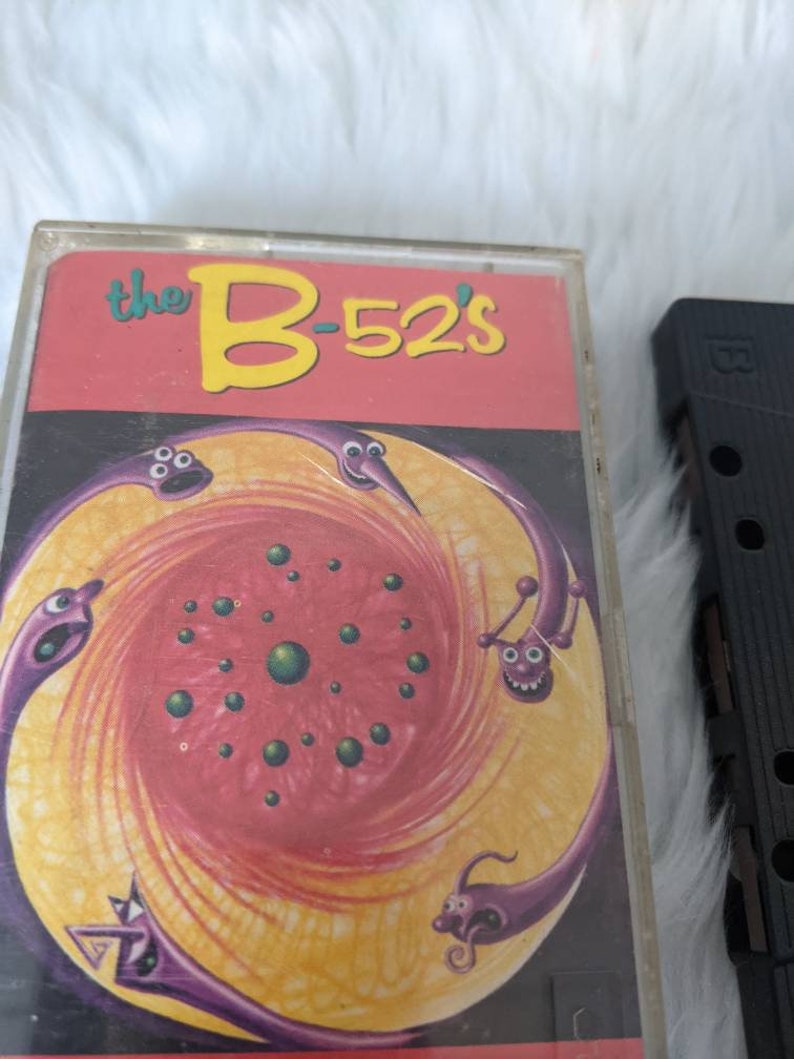 May include: A cassette tape featuring a red, yellow and purple illustration of a swirling galaxy with cartoon worm-like creatures. The text "the B-52's" is printed in yellow on a red background.