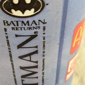 May include: A blue and white cup with the Batman Returns logo. The logo features the Batman symbol and the text "Batman Returns". The text "&copy; 1992" and "&copy; DC Comics Inc." are also visible.