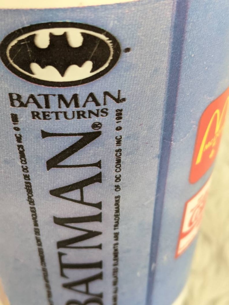 May include: A blue and white cup with the Batman Returns logo. The logo features the Batman symbol and the text "Batman Returns". The text "&copy; 1992" and "&copy; DC Comics Inc." are also visible.