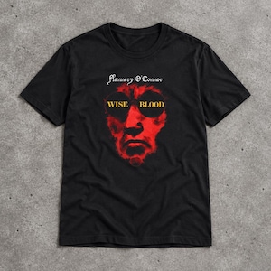 Könnte beinhalten: Ein schwarzes Kurzarm-T-Shirt mit einem Grafikdruck eines roten Gesichts, das eine runde dunkle Brille trägt. Der Text Flannery O'Connor steht in Weiß über dem Gesicht und die Wörter WISE BLOOD sind in Gelb auf den Brillengläsern aufgedruckt.