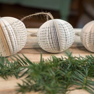 May include: Three white paper ornaments with pages from a book folded into a sphere shape. The ornaments are hanging from twine and are resting on a wooden surface with green pine branches.