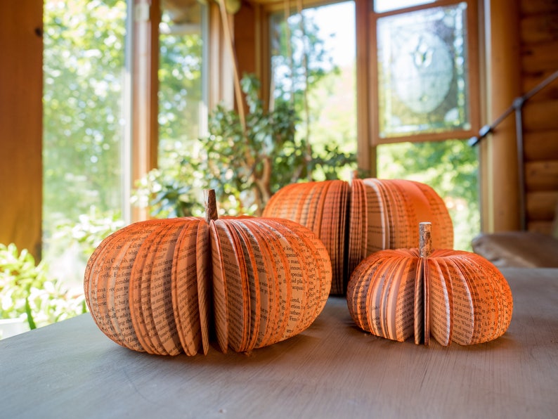 Op de afbeelding: Drie decoratieve pompoenen gemaakt van gevouwen boekpagina's. De pompoenen zijn oranje en hebben een rustieke, handgemaakte uitstraling.