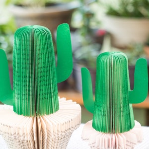 Op de afbeelding: Twee groene papieren cactussen gemaakt van gevouwen boekpagina's. De cactussen staan op een wit oppervlak.