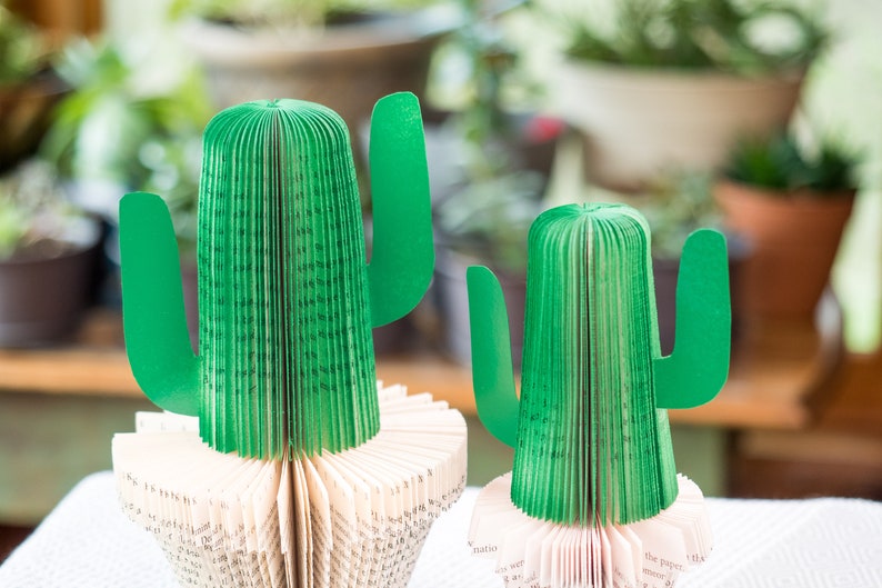Op de afbeelding: Twee groene papieren cactussen gemaakt van gevouwen boekpagina's. De cactussen staan op een wit oppervlak.