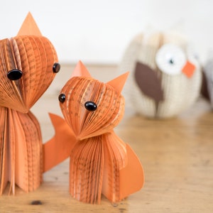 May include: Two orange paper foxes made from book pages with black eyes. The foxes are standing on a wooden surface. Two owls made from book pages are in the background.