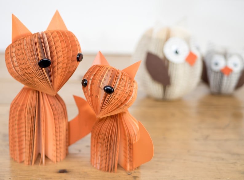 May include: Two orange paper foxes made from book pages with black eyes. The foxes are standing on a wooden surface. Two owls made from book pages are in the background.