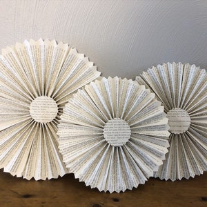 May include: Three paper fan decorations made from the pages of old books. The fans are white with black text and are arranged in a semi-circle on a wooden surface.