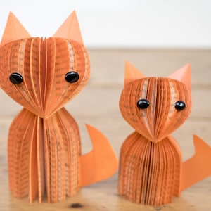 May include: Two orange paper fox sculptures made from book pages with black button eyes. The foxes are standing on a wooden surface.