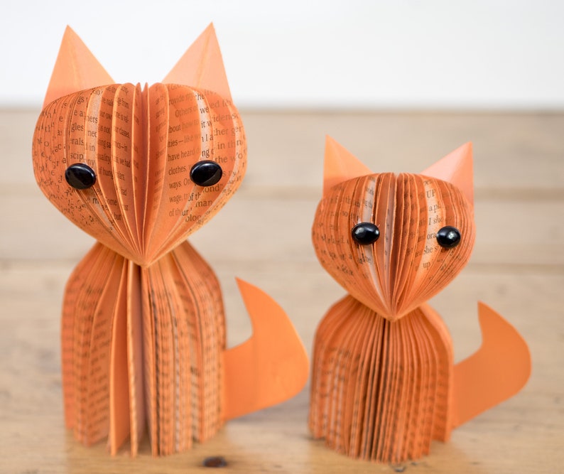 May include: Two orange paper fox sculptures made from book pages with black button eyes. The foxes are standing on a wooden surface.