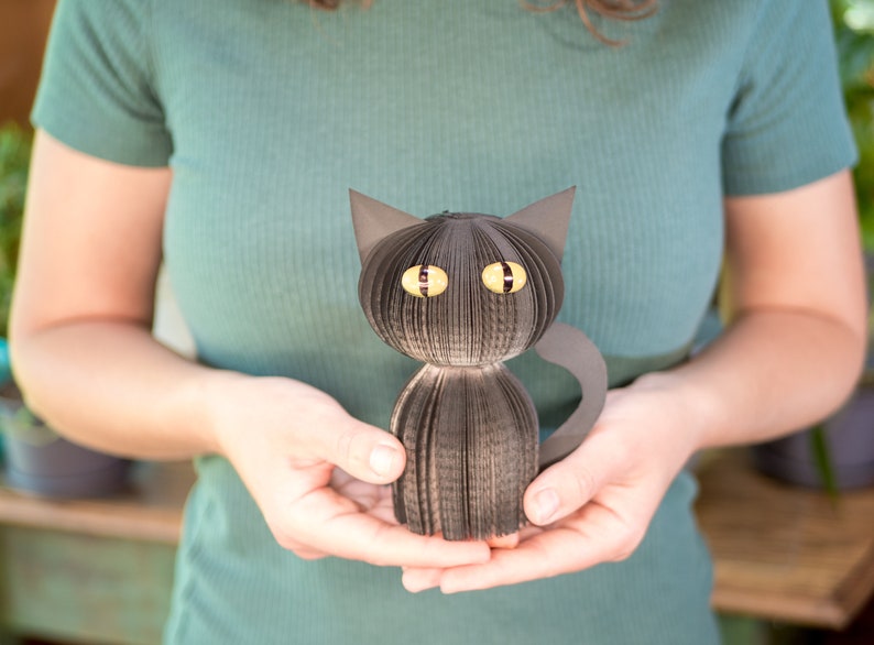 May include: A black paper cat with yellow eyes is being held by a person. The cat is made of folded paper and has a tail that curls around its body.