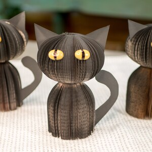 May include: Three black paper cat sculptures with yellow eyes. The cats are made from folded pages of a book.