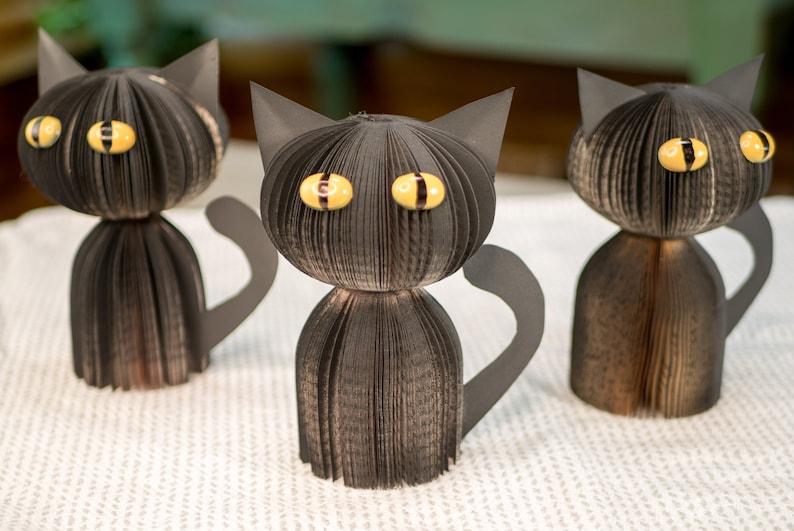 May include: Three black paper cat sculptures with yellow eyes. The cats are made from folded pages of a book.