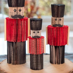 May include: Three book page nutcracker figurines, made from folded pages of books, standing on a wooden slice. The nutcrackers are black, red, and white with black mustaches and eyes.