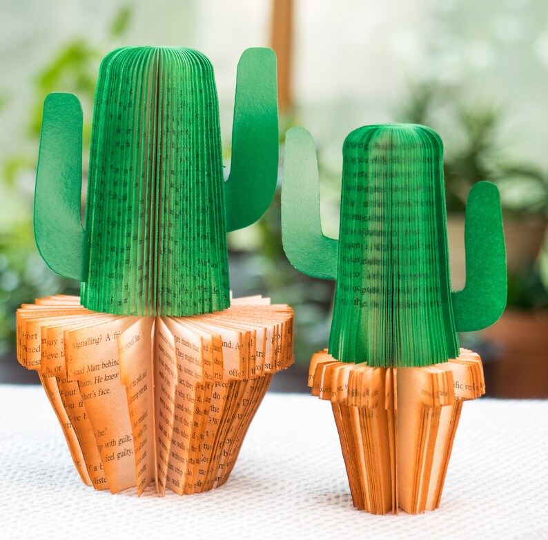 Op de afbeelding: Twee groene papieren cactus-boekenleggers gemaakt van gevouwen boekpagina's. De boekenleggers staan op een wit oppervlak.