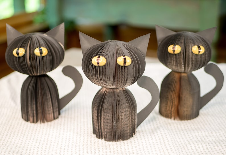 May include: Three black paper cat sculptures with yellow eyes. The cats are made from folded pages of a book and have a 3D effect.