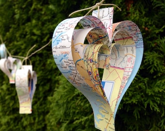 May include: A handmade heart-shaped ornament crafted from repurposed road maps. The ornament is suspended by a natural twine string, showcasing layers of colorful map details. Additional heart-shaped ornaments are visible in the background, creating a decorative display.