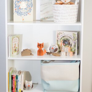 May include: A white bookshelf with five shelves. The top shelf has a sign that says "All of God's grace in one tiny face". The second shelf has a book and a basket with the word "mother" repeated in different languages. The third shelf has a book, a hedgehog, a fox, an owl, and blocks that spell "G, S, J, Y". The fourth shelf has a stack of books and an owl. The bottom shelf has a book, a toy, and a wooden toy with a fox on it.