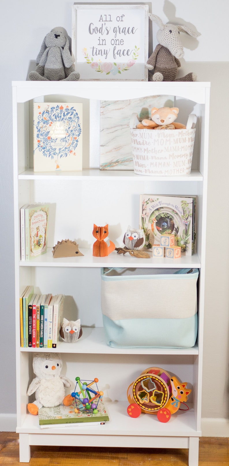 May include: A white bookshelf with five shelves. The top shelf has a sign that says "All of God's grace in one tiny face". The second shelf has a book and a basket with the word "mother" repeated in different languages. The third shelf has a book, a hedgehog, a fox, an owl, and blocks that spell "G, S, J, Y". The fourth shelf has a stack of books and an owl. The bottom shelf has a book, a toy, and a wooden toy with a fox on it.