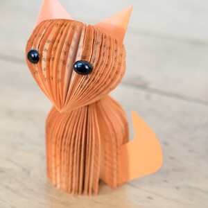 May include: A handmade orange paper cat figurine with black eyes. The cat is made from folded pages of a book.