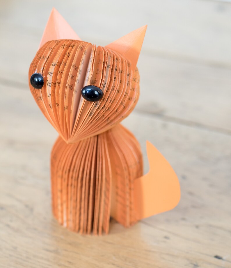 May include: A handmade orange paper cat figurine with black eyes. The cat is made from folded pages of a book.