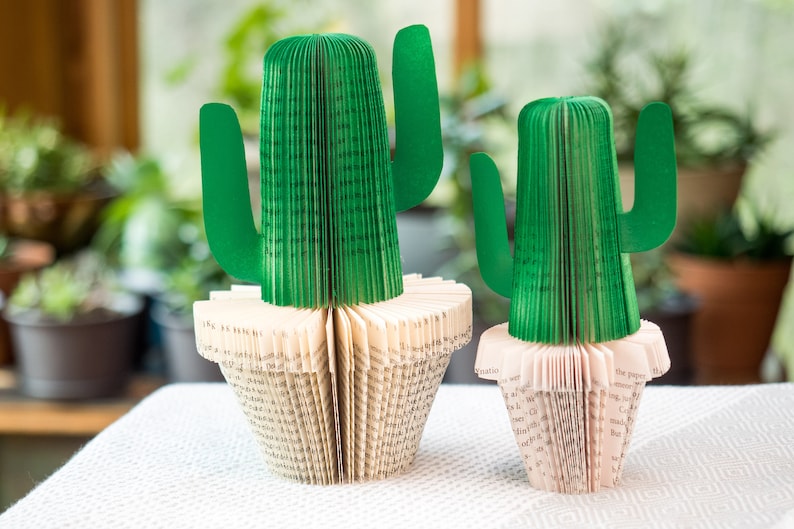 Op de afbeelding: Twee groene papieren cactussen in witte potten gemaakt van gevouwen boekpagina's. De cactussen staan op een wit tafelkleed.