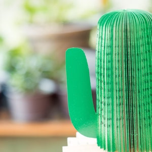 Op de afbeelding: Een groene papieren cactus met een boekrugontwerp. De cactus is gemaakt van gevouwen pagina's en heeft een groene papieren arm.