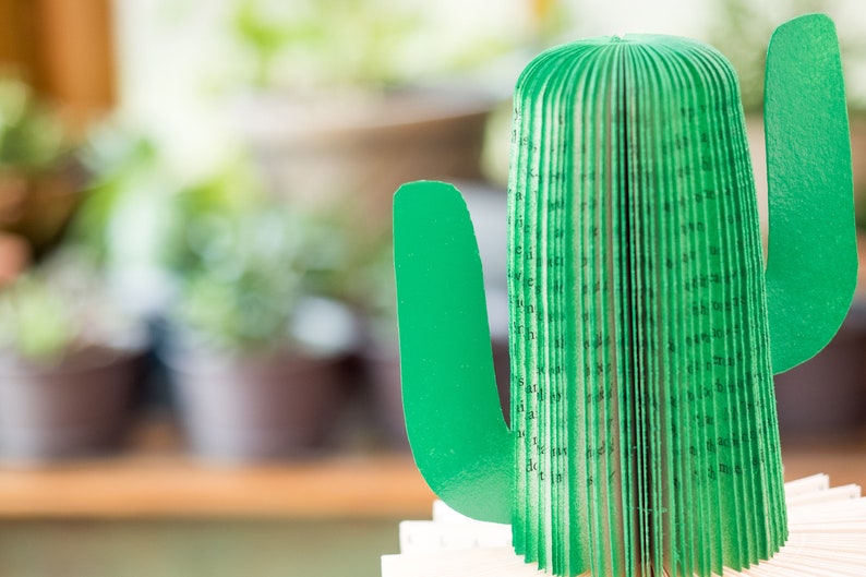 Op de afbeelding: Een groene papieren cactus met een boekrugontwerp. De cactus is gemaakt van gevouwen pagina's en heeft een groene papieren arm.