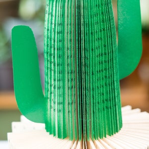 Op de afbeelding: Een groene papieren cactus gemaakt van de pagina's van een boek. De cactus is gevouwen tot een driedimensionale vorm met een lang, slank lichaam en twee armen. De pagina's van het boek zijn zichtbaar aan de buitenkant van de cactus, en de tekst is in zwart gedrukt.