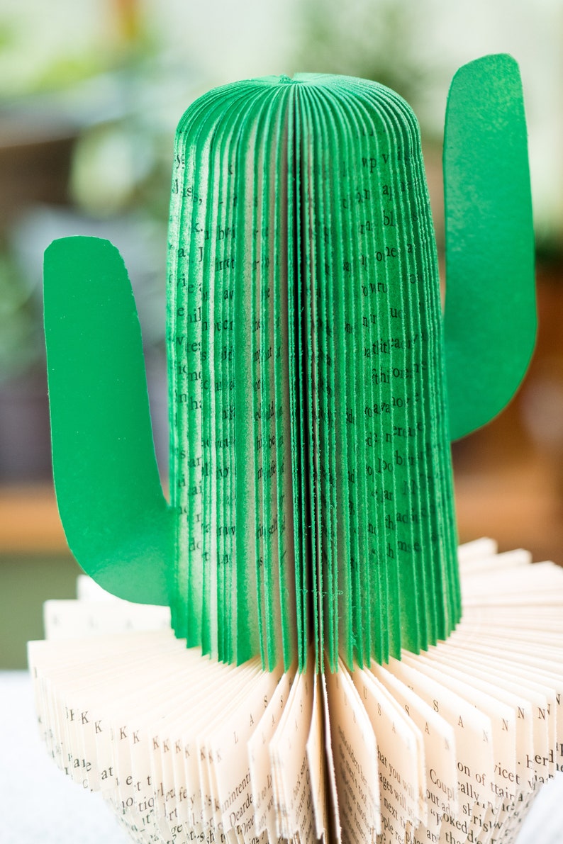 Op de afbeelding: Een groene papieren cactus gemaakt van de pagina's van een boek. De cactus is gevouwen tot een driedimensionale vorm met een lang, slank lichaam en twee armen. De pagina's van het boek zijn zichtbaar aan de buitenkant van de cactus, en de tekst is in zwart gedrukt.