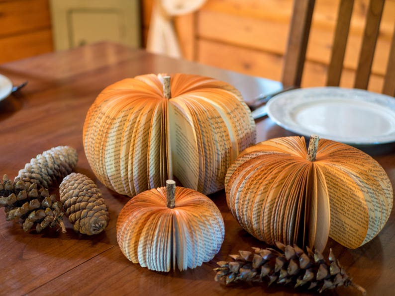 Op de afbeelding: Drie boekpagina-pompoenen op een houten tafel met dennenappels. De pompoenen zijn gemaakt van boekpagina's en zijn gevouwen in de vorm van een pompoen. De dennenappels zijn bruin en hebben een natuurlijke afwerking.