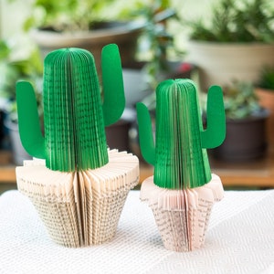 Op de afbeelding: Twee groene papieren cactussculpturen gemaakt van gevouwen boekpagina's. De cactussculpturen staan op een wit tafelkleed.