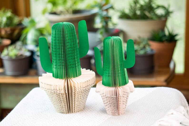 Op de afbeelding: Twee groene papieren cactussculpturen gemaakt van gevouwen boekpagina's. De cactussculpturen staan op een wit tafelkleed.