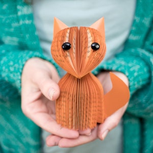 May include: A hand-crafted fox figurine made from folded pages of a book. The fox is orange and has black eyes. The figurine is being held in the hands of a person wearing a teal sweater.
