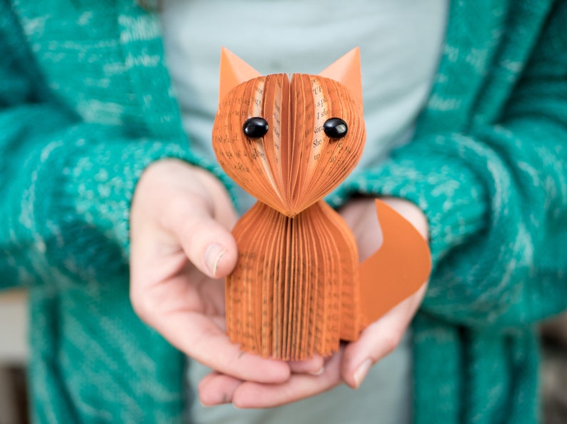 May include: A hand-crafted fox figurine made from folded pages of a book. The fox is orange and has black eyes. The figurine is being held in the hands of a person wearing a teal sweater.