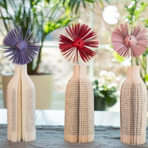 May include: Three decorative vases made from folded book pages, each holding a paper flower. The flowers are in shades of purple, red, and pink. The vases are arranged on a gray surface in front of a window with greenery.