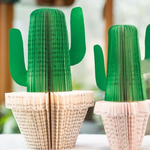 Op de afbeelding: Twee groene papieren cactussen in witte potten. De cactussen zijn gemaakt van gevouwen boekpagina's. De grotere cactus is groter dan de kleinere cactus.