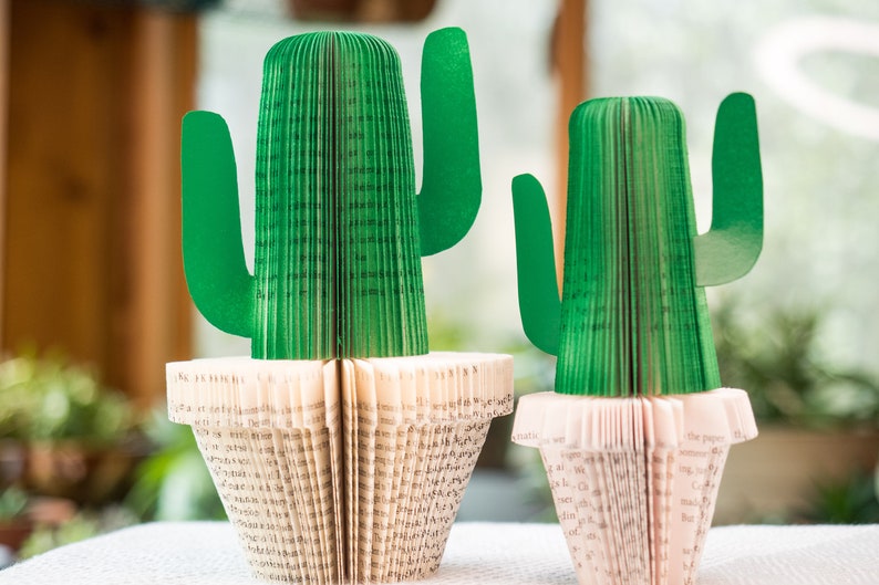 Op de afbeelding: Twee groene papieren cactussen in witte potten. De cactussen zijn gemaakt van gevouwen boekpagina's. De grotere cactus is groter dan de kleinere cactus.