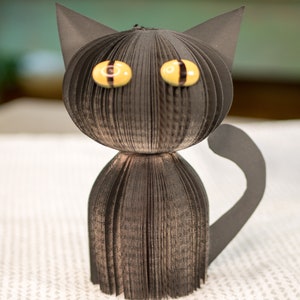 May include: A black cat made from folded pages of a book. The cat has yellow eyes with black pupils and a black tail.