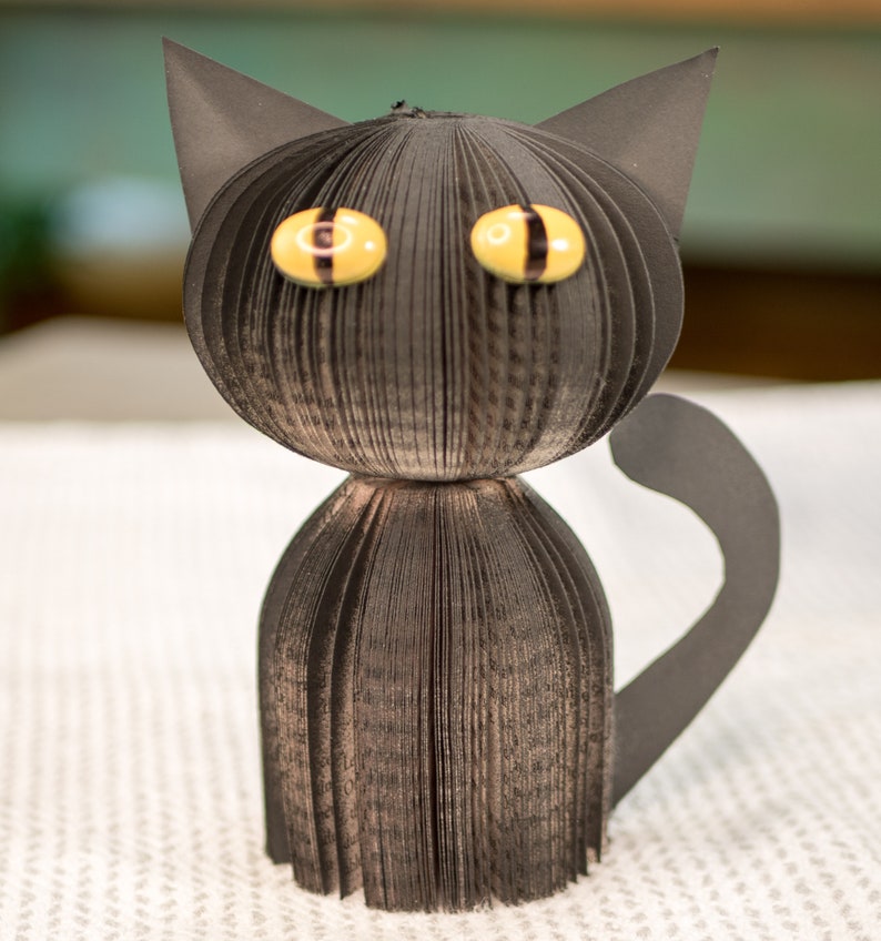 May include: A black cat made from folded pages of a book. The cat has yellow eyes with black pupils and a black tail.