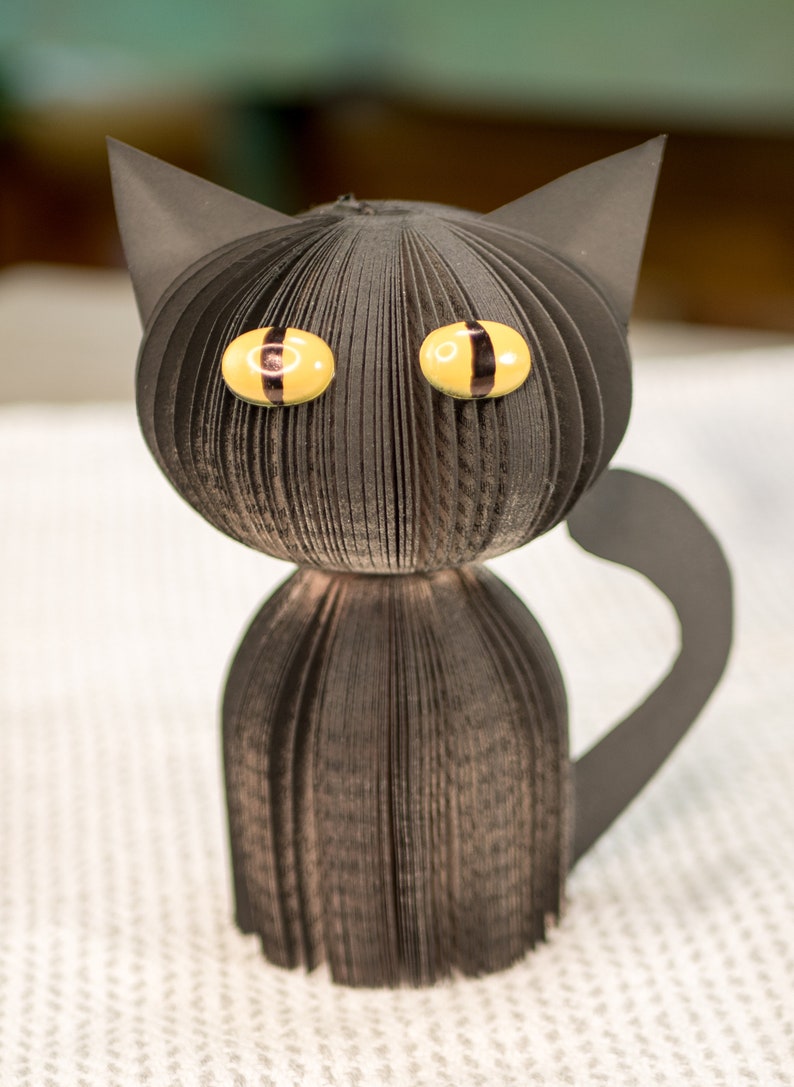 May include: A black paper cat with yellow eyes and a black tail. The cat is made from folded paper and has a 3D effect.
