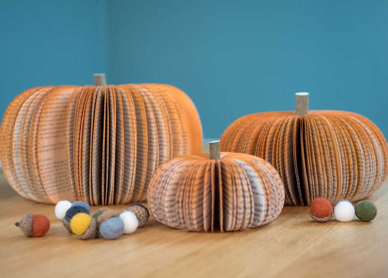 Op de afbeelding: Drie oranje papieren pompoenen gemaakt van boekpagina's, met een steel op elk. De pompoenen zijn gerangschikt op een houten oppervlak met kleine, kleurrijke viltballetjes die eromheen zijn verspreid.