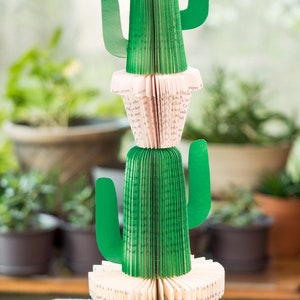 Op de afbeelding: Een groene papieren cactus met een roze papieren basis. De cactus is gemaakt van de pagina's van een boek en heeft drie armen. De cactus staat op een wit tafelkleed.