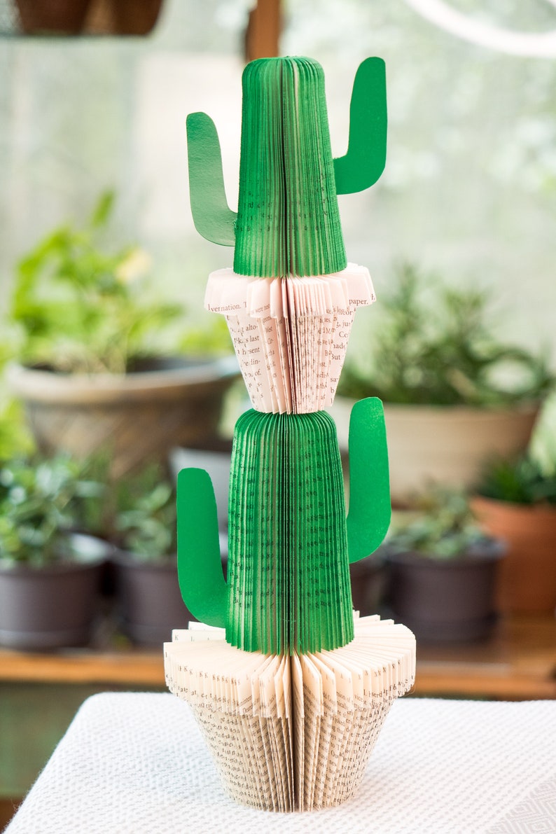 Op de afbeelding: Een groene papieren cactus met een roze papieren basis. De cactus is gemaakt van de pagina's van een boek en heeft drie armen. De cactus staat op een wit tafelkleed.